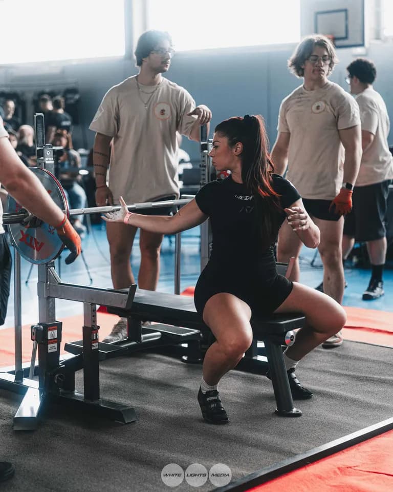 Une athlète assise sur le banc tend le bras vers la barre pour s'installer avant un développé couché, entourée de pareurs. Photo @melanie__immo / @whitelightsmediafr.