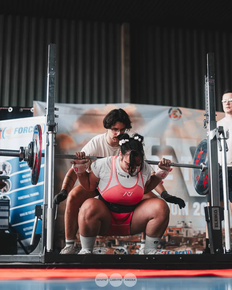 Une athlète en singlet rose descend en squat profond, un pareur juste derrière elle, sur un plateau FFForce. Photo @dumascamille_ / @whitelightsmediafr.