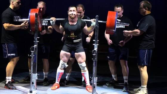 Athlète en position de départ au squat, barre chargée sur le dos, entouré de pareurs. Image du programme Smolov Jr de l’app Sarox.